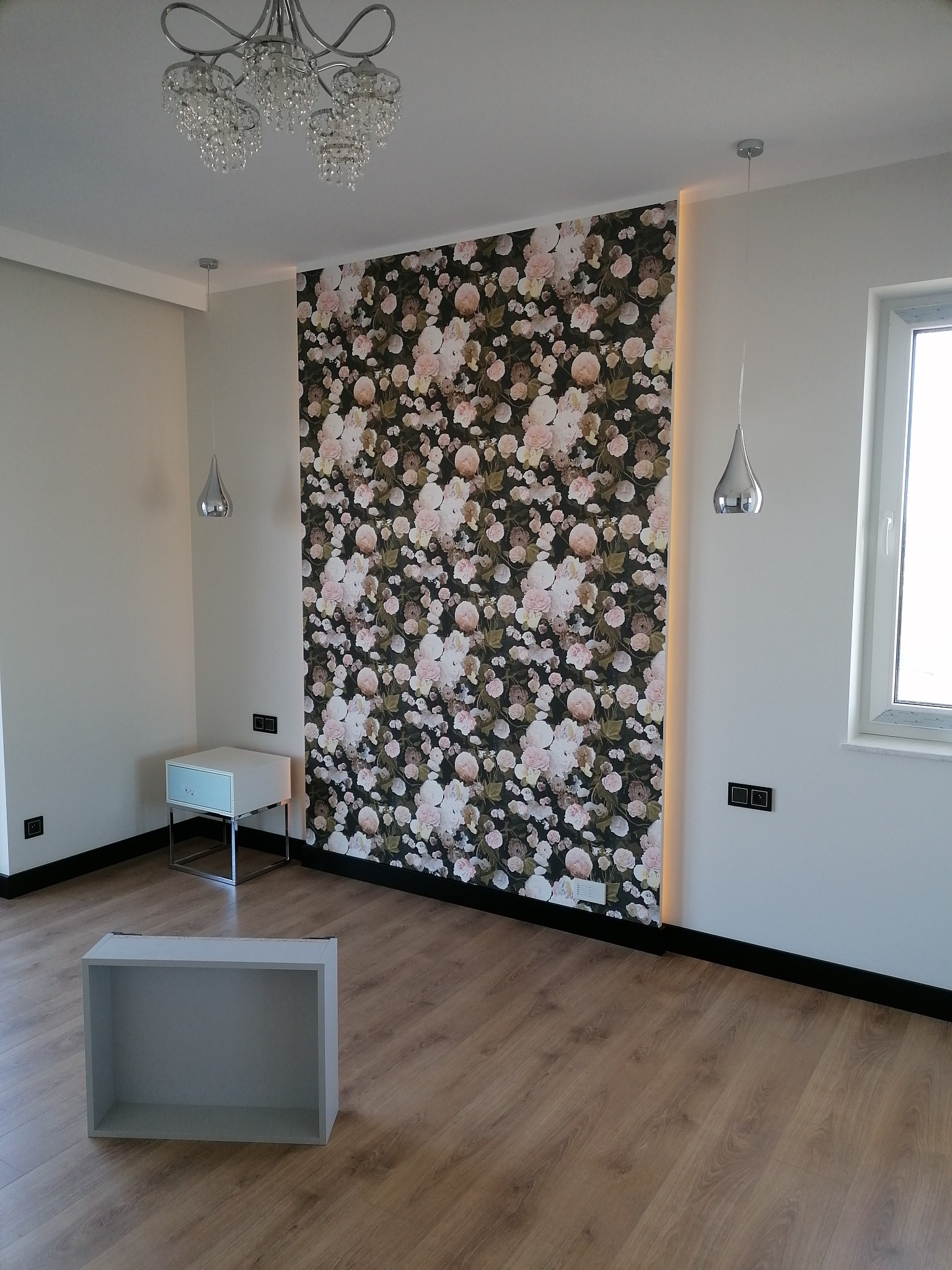 Pokój po remoncie z tapetą w kwiaty na jednej ze ścian, drewnianą podłogą, nowoczesnym oświetleniem i niedokończonym meblem na podłodze.