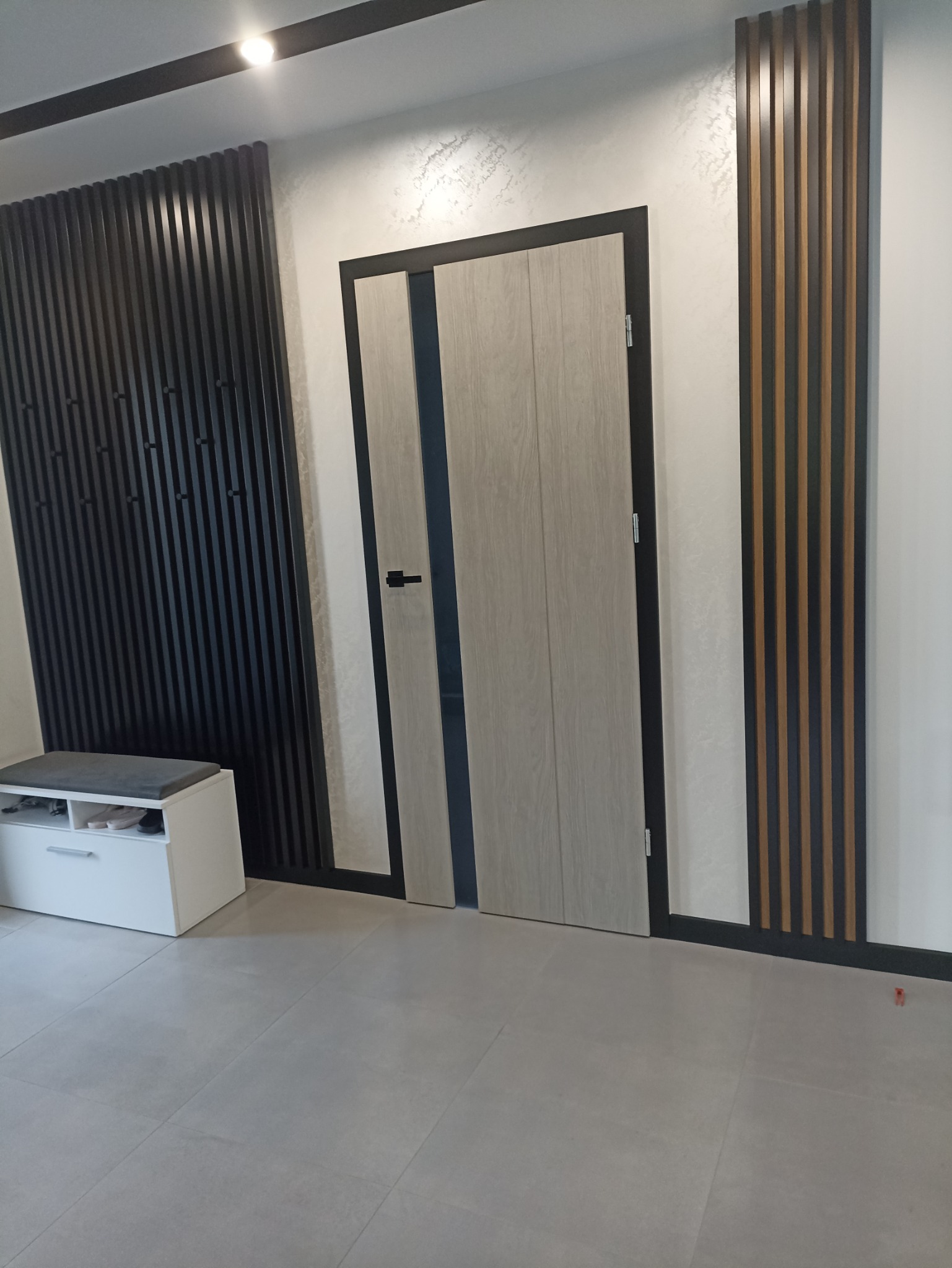 Nowoczesne wnętrze: drewniane drzwi wewnętrzne z czarną ościeżnicą, dekoracyjne lamele ścienne w kolorze czarnym i brązowym, biała komoda z szarą poduszką na siedzisku, szara podłoga z płytek.