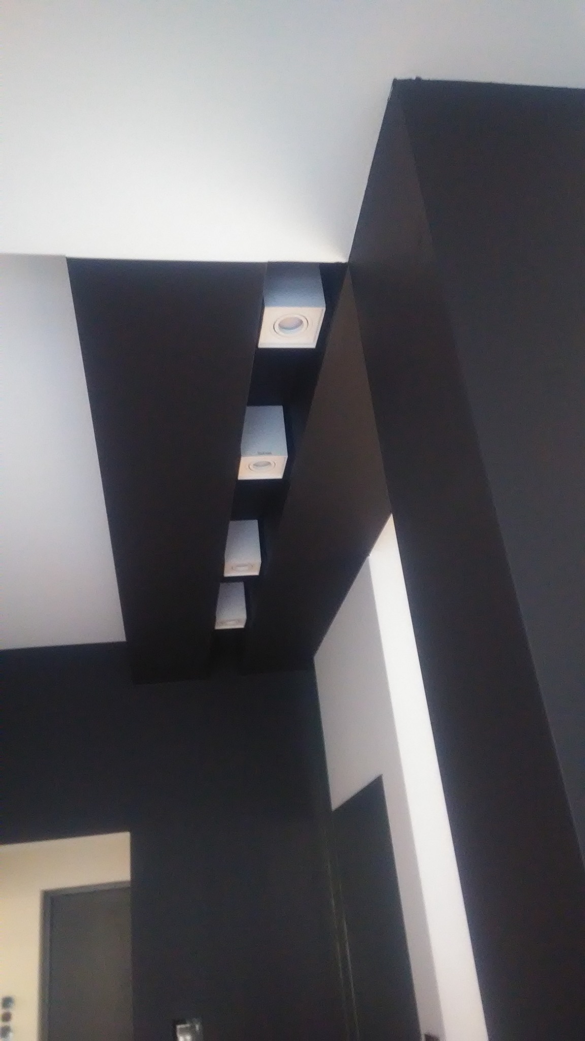 Czarno-białe wnętrze w Łukowie z rzędem czterech kwadratowych, wpuszczanych lamp sufitowych w czarnej wnęce, widok z dołu.