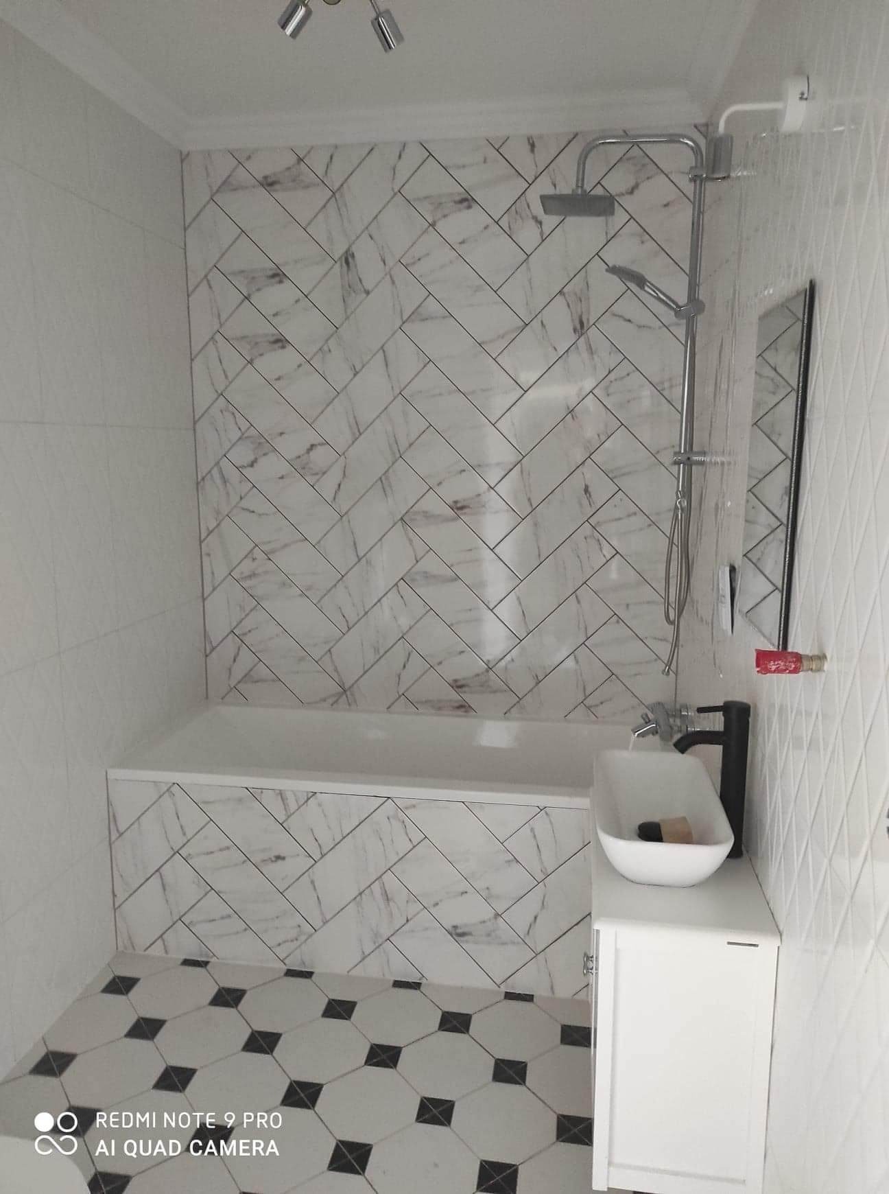 Wykończona łazienka z biało-szarą marmurkową glazurą ułożoną w jodełkę na ścianie nad wanną, białą umywalką nablatową z czarną baterią i podłogą w biało-czarne płytki o geometrycznym wzorze.