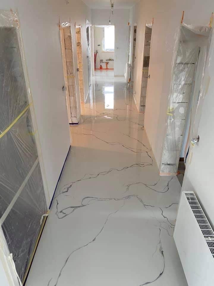 Korytarz po remoncie z nową, błyszczącą podłogą imitującą marmur, drzwi zabezpieczone folią malarską, widoczny grzejnik i dalsze pomieszczenia w perspektywie.