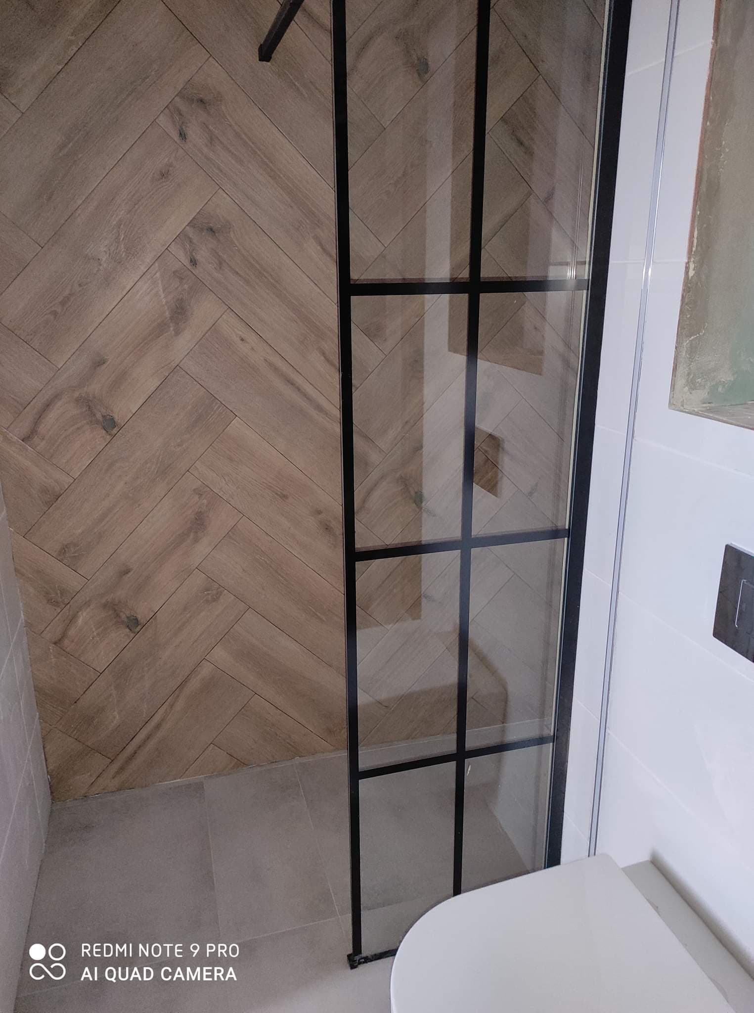 Wykończona łazienka z kabiną prysznicową typu walk-in z czarnymi szprosami, drewnopodobne płytki ułożone w jodełkę na ścianie, szare płytki podłogowe, biała ceramika sanitarna.