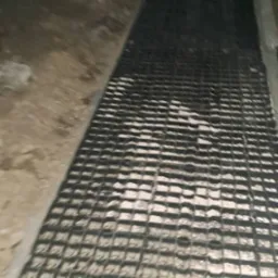 Ułożona krata drenażowa na podłożu z betonu, widoczne fragmenty gruntu i betonowej ściany w słabym oświetleniu.