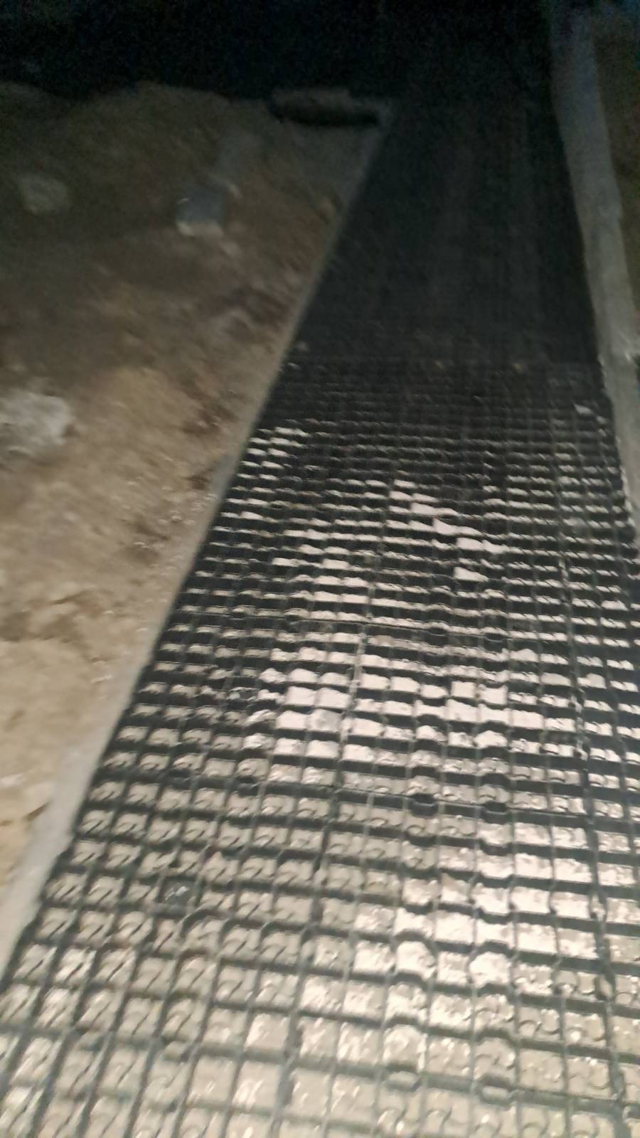 Ułożona krata drenażowa na podłożu z betonu, widoczne fragmenty gruntu i betonowej ściany w słabym oświetleniu.
