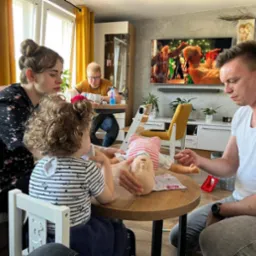 Instruktor demonstruje na manekinie niemowlęcia resuscytację krążeniowo-oddechową w obecności kobiety i dziecka, w tle mężczyzna przy stole i telewizor.