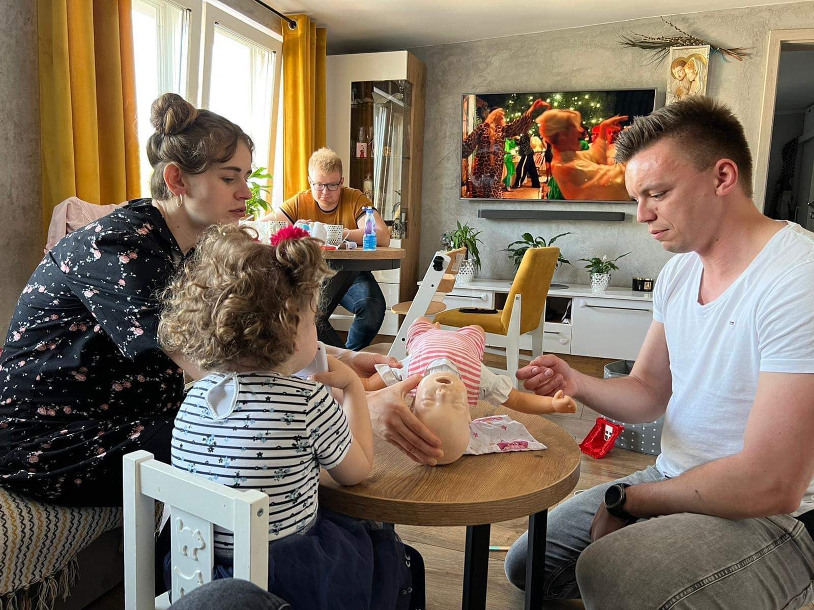 Instruktor demonstruje na manekinie niemowlęcia resuscytację krążeniowo-oddechową w obecności kobiety i dziecka, w tle mężczyzna przy stole i telewizor.