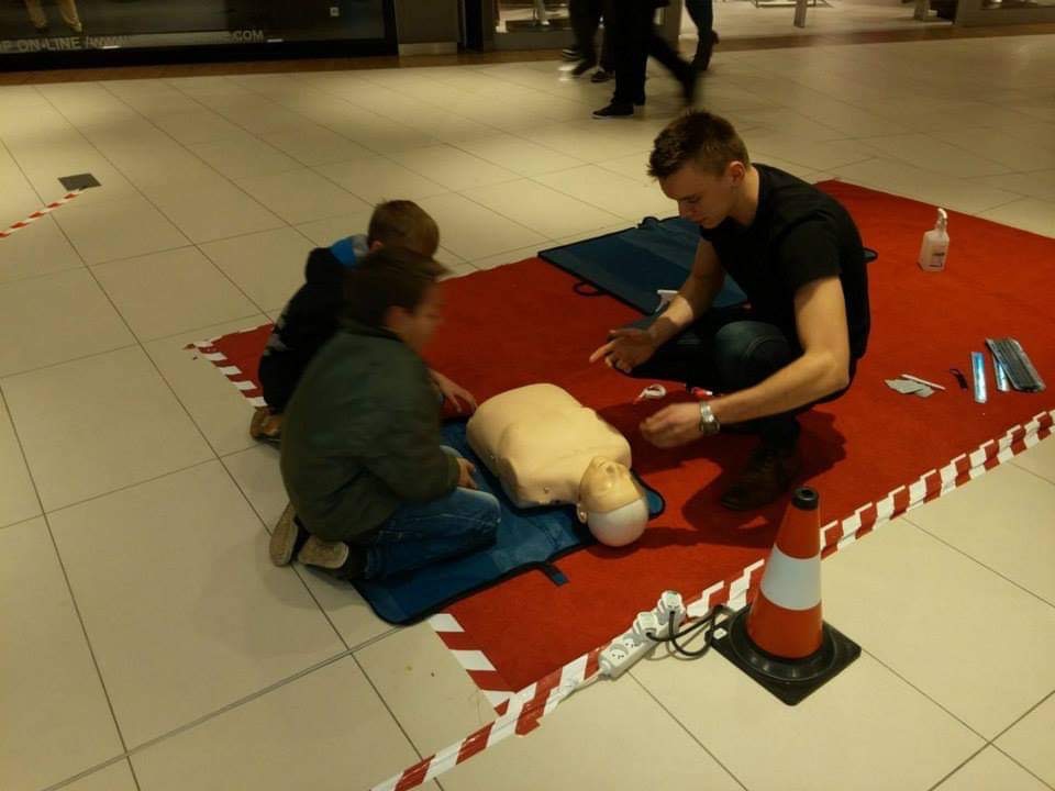 Instruktor demonstruje resuscytację krążeniowo-oddechową na manekinie przed dwójką dzieci na czerwonym dywanie w jasnym, otwartym pomieszczeniu.