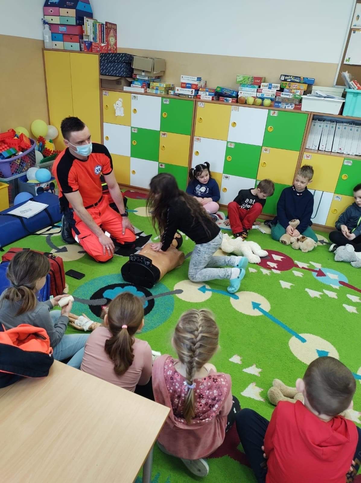 Instruktor w pomarańczowym uniformie i masce demonstruje resuscytację na manekinie przed grupą dzieci siedzących na zielonym dywanie w sali lekcyjnej.