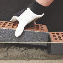 Murarz w rękawicach układa cegłę z szarą krawędzią na warstwie zaprawy, widoczna linka murarska.