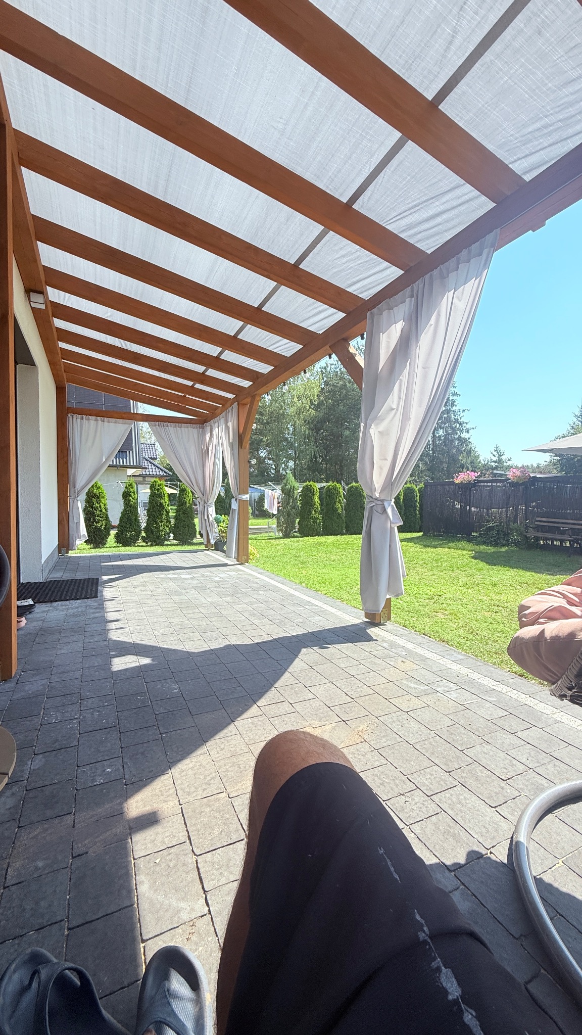 Zadaszony taras z drewnianą konstrukcją i zwiewnymi, białymi zasłonami. Widok na zadbany ogród z trawnikiem i tujami. Cień na kostce brukowej.