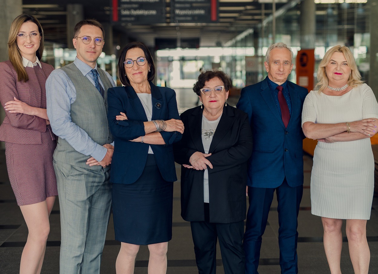 Zespół biura rachunkowego pozuje z założonymi rękami na tle nowoczesnego biurowca z przeszkloną fasadą, widoczne tablice informacyjne.