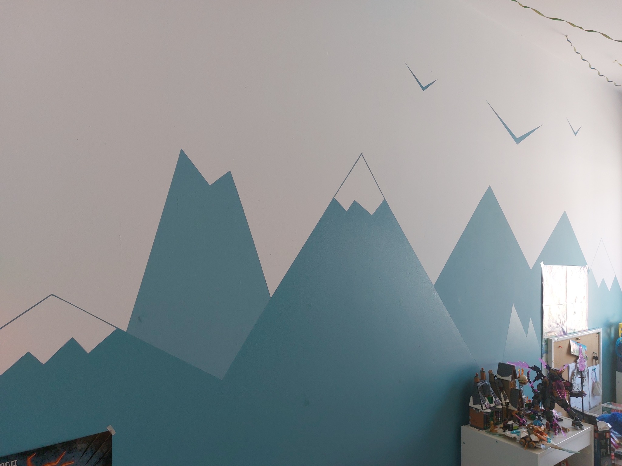 Ściana pokoju dziecięcego z ręcznie malowanym motywem górskim w odcieniach bieli i błękitu, uzupełniona o sylwetki ptaków w locie, z zabawkami na pierwszym planie.
