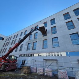 FIRMA REMONTOWO-WYKOŃCZENIOWA BORKOWSKI ŁUKASZ - Montaż paneli elewacyjnych na budynku z użyciem podnośnika teleskopowego. Widoczne rusztowania i materiały budowlane. Dzień, błękitne niebo.