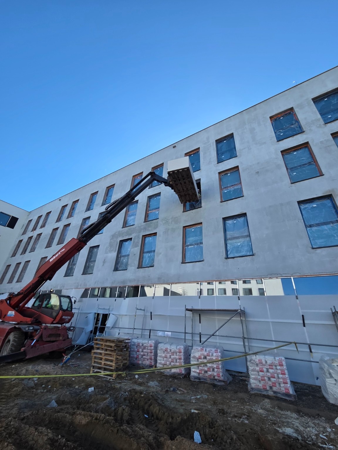 Montaż paneli elewacyjnych na budynku z użyciem podnośnika teleskopowego. Widoczne rusztowania i materiały budowlane. Dzień, błękitne niebo.