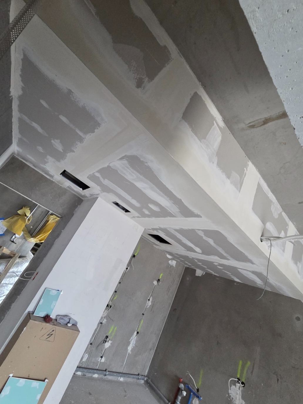 Sufit podwieszany z płyt gipsowo-kartonowych w trakcie szpachlowania, widoczne otwory wentylacyjne i instalacje elektryczne na ścianach. Surowy stan wykończenia.