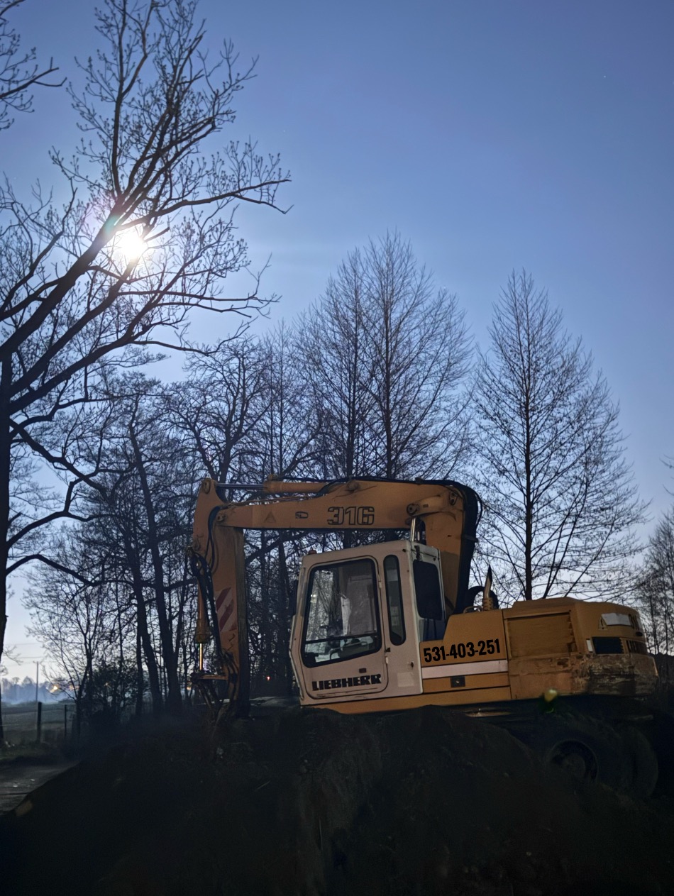 Żółta koparka Liebherr 316 na tle zimowego krajobrazu oświetlonego księżycem, widoczna z boku, z numerem telefonu na kabinie.