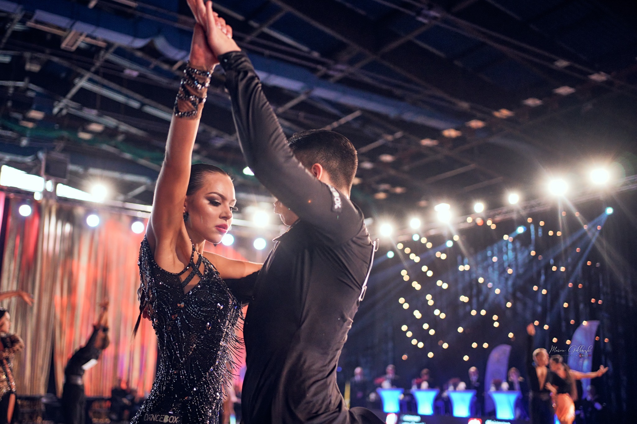 Para tancerzy w trakcie występu tanecznego; kobieta w czarnej, błyszczącej sukience z frędzlami, mężczyzna w czarnym stroju; w tle oświetlenie sceniczne.