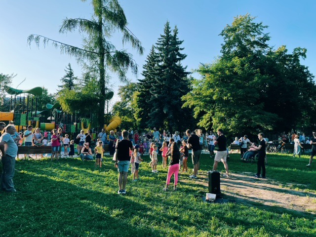 Lekcja tańca na świeżym powietrzu, grupa ludzi w różnym wieku tańczy na trawie w parku, w tle plac zabaw i drzewa.