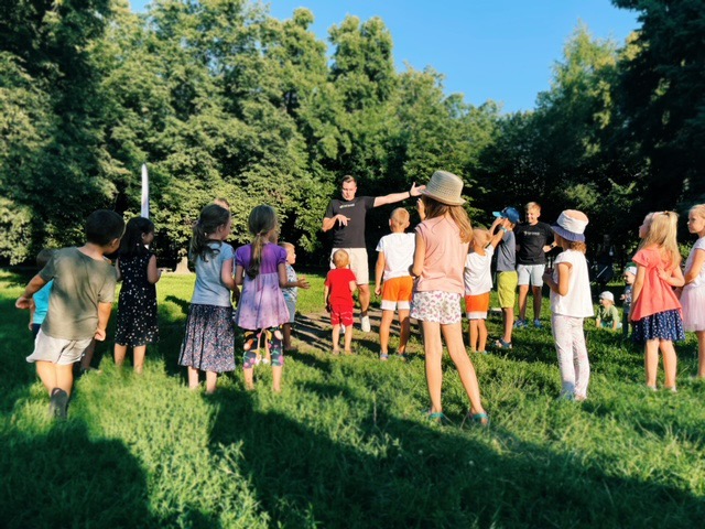 Grupa dzieci słucha instruktora na trawie w parku, prawdopodobnie podczas zajęć tanecznych na świeżym powietrzu; w tle drzewa i fontanna.