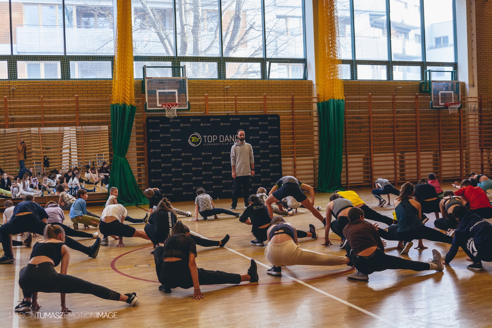 Grupa osób rozciąga się na parkiecie sali gimnastycznej przed czarnym banerem z logo 'Top Dance', pod nadzorem instruktora.
