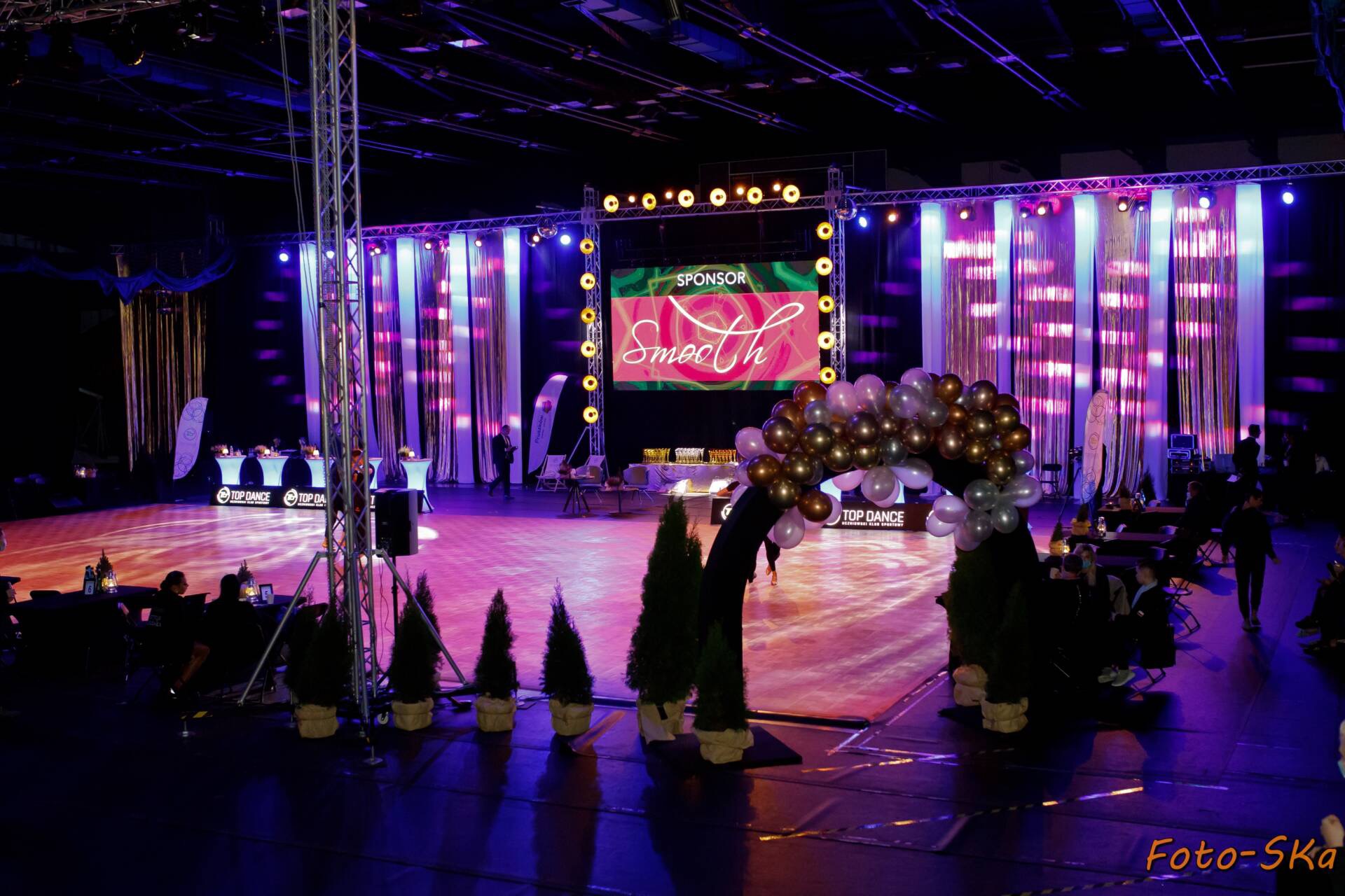 Widok na salę taneczną podczas wydarzenia, z dekoracjami świetlnymi, balonami i roślinami, widoczny parkiet i stoliki dla gości.