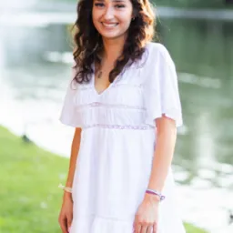 Uśmiechnięta młoda kobieta w białej sukience i wianku z białych kwiatów na głowie, pozująca na tle jeziora w parku.