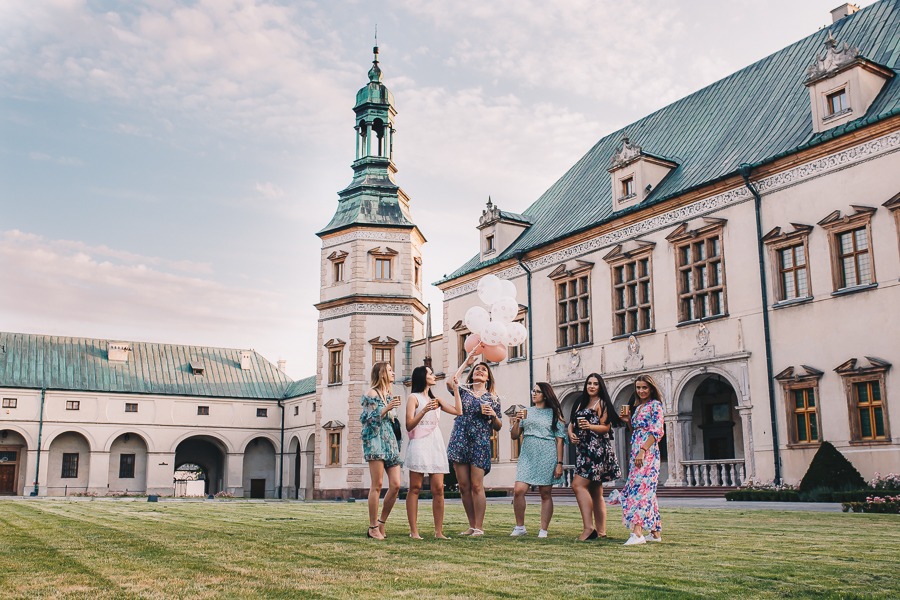 Grupa siedmiu kobiet świętuje z balonami i napojami na trawniku przed zabytkowym pałacem z wieżą i arkadami.
