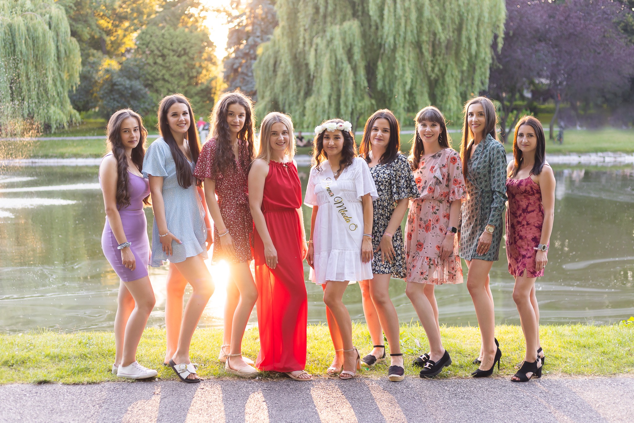 Grupa dziewięciu kobiet w różnorodnych sukienkach pozuje na tle jeziora i zieleni; jedna z nich ma szarfę z napisem 'Przyszła Młoda' i wianek na głowie, sugerując wieczór panieński.