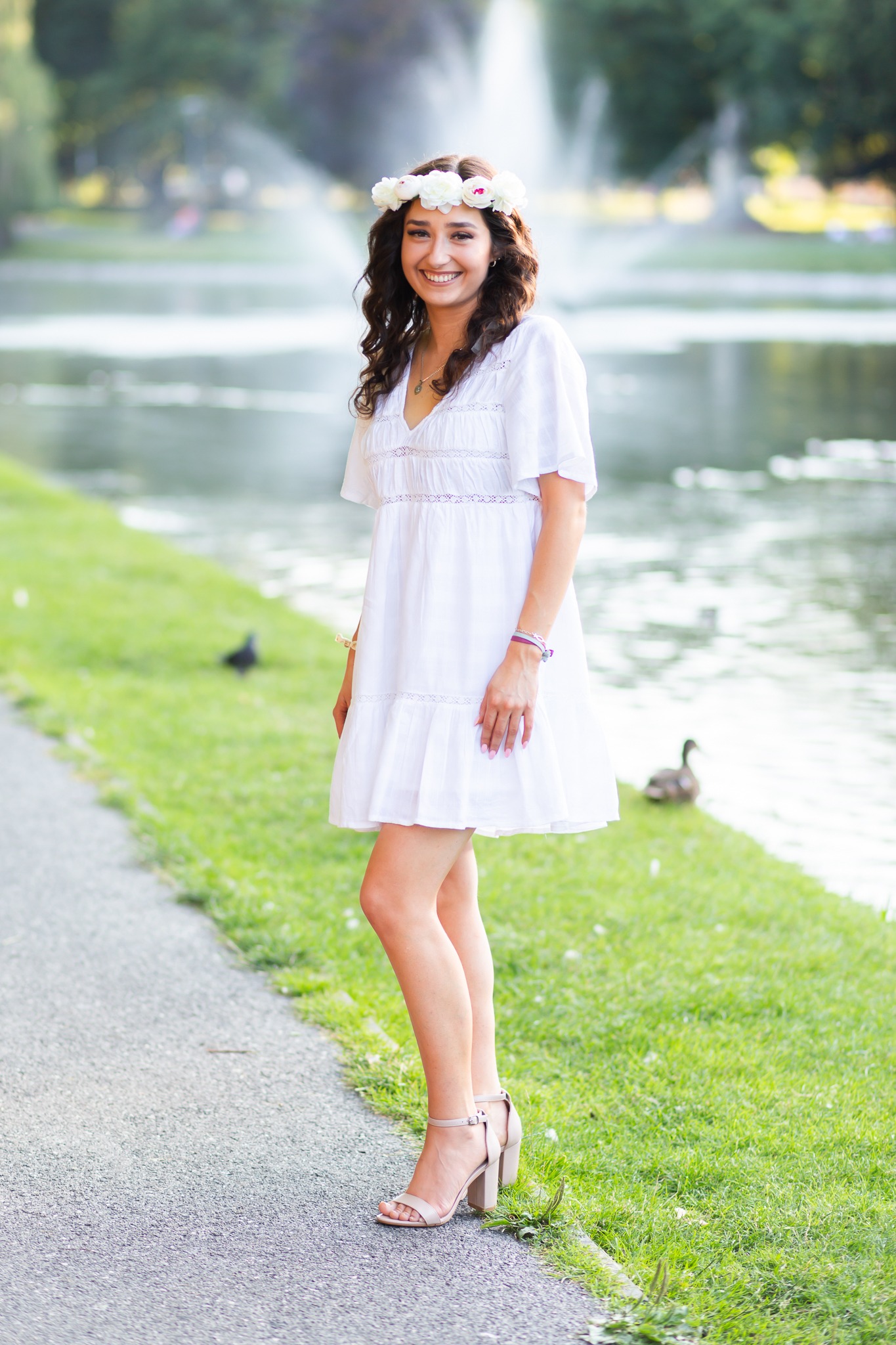 Uśmiechnięta młoda kobieta w białej sukience i wianku na głowie, pozująca na tle parku z fontanną.
