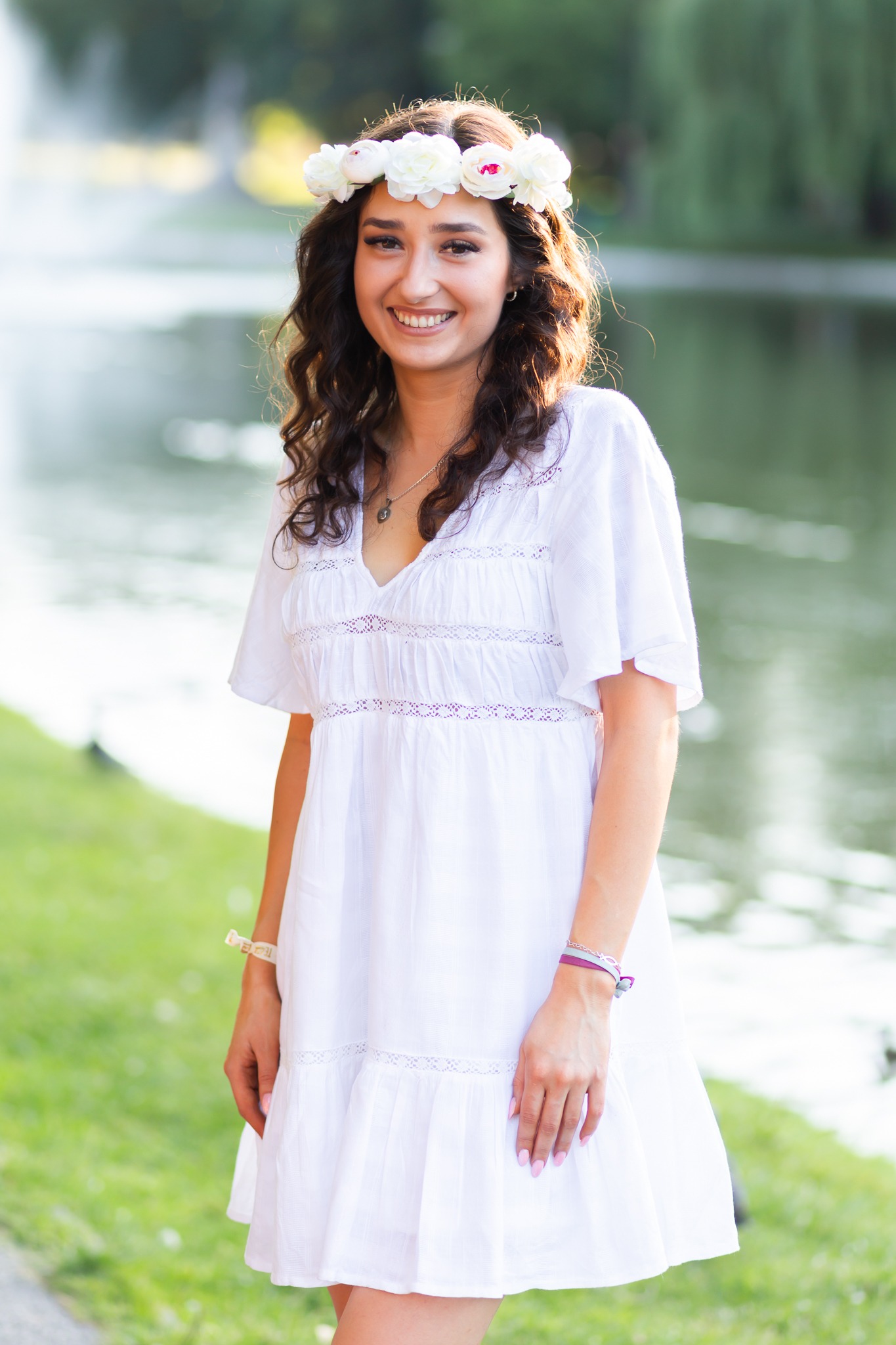 Uśmiechnięta młoda kobieta w białej sukience i wianku z białych kwiatów na głowie, pozująca na tle jeziora w parku.