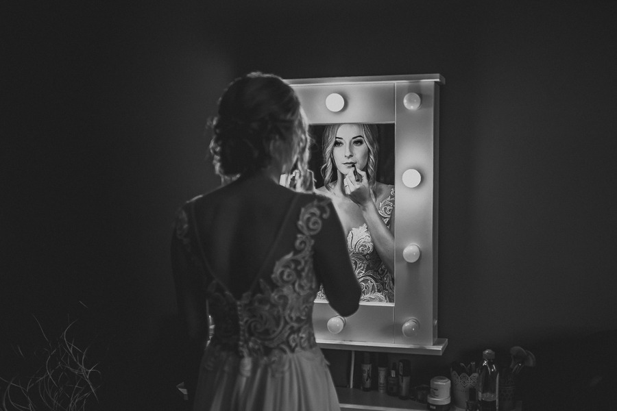 Czarno-białe zdjęcie panny młodej, widzianej od tyłu, poprawiającej makijaż ust przed lustrem z oświetleniem, na którym widoczne jest jej odbicie.