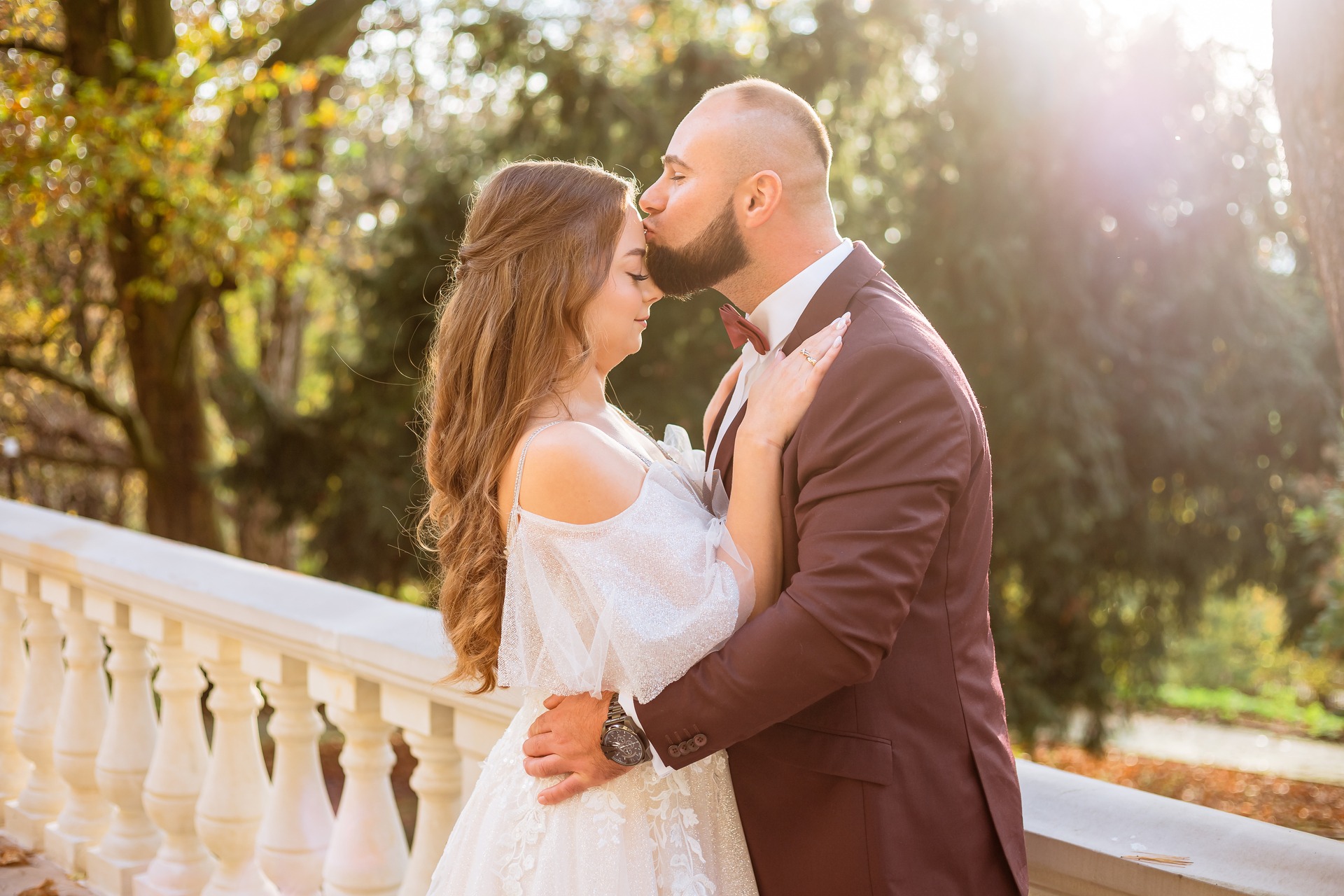 Para młoda w objęciach na tle parku w słoneczny dzień, panna młoda w białej sukni z odkrytymi ramionami, pan młody w bordowym garniturze i muszce, pocałunek w czoło.