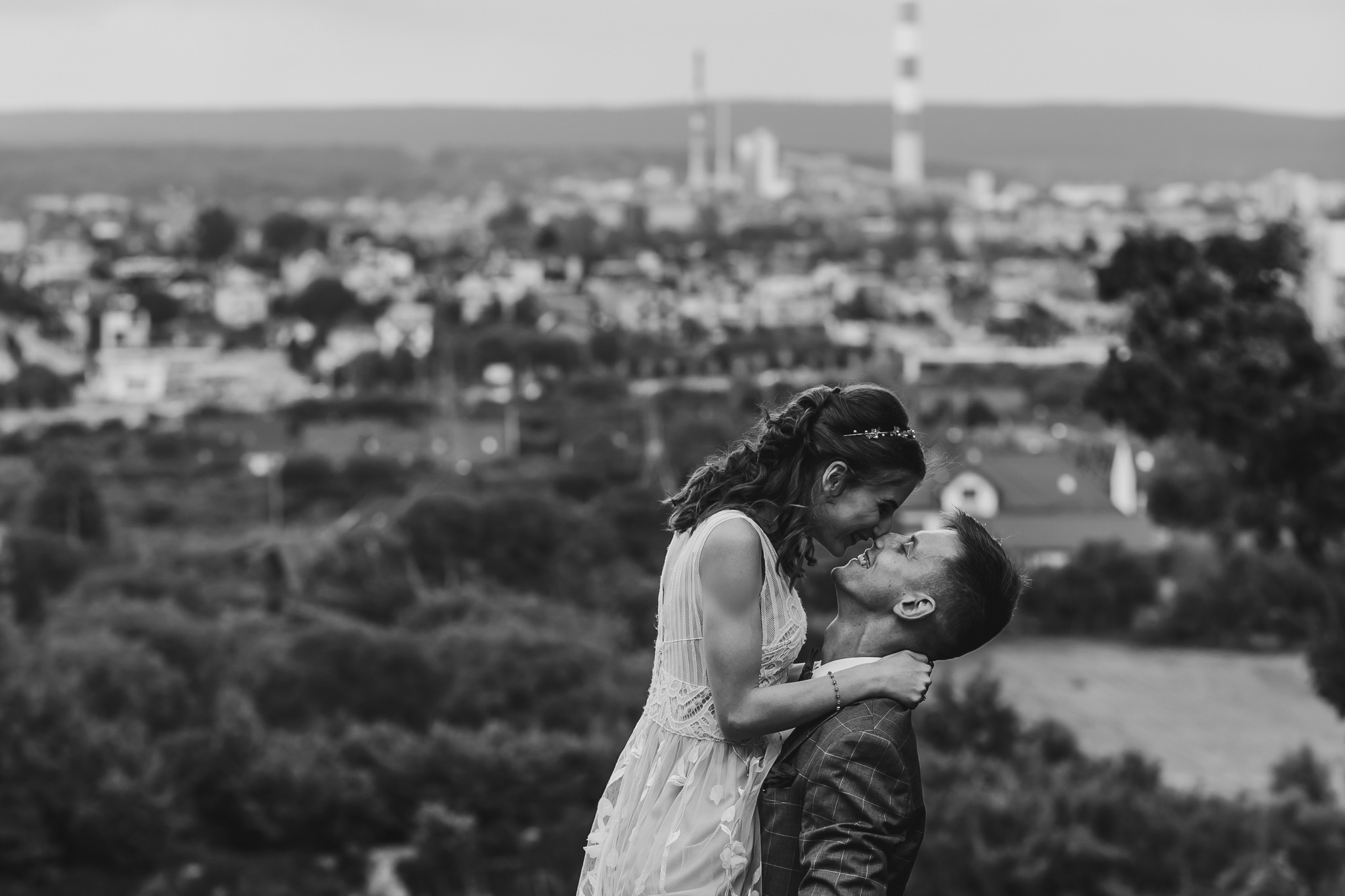 Czarno-białe zdjęcie pary młodej, panna młoda w koronkowej sukni i tiarze, obejmuje pana młodego w kraciastej marynarce na tle miasta i komina przemysłowego.