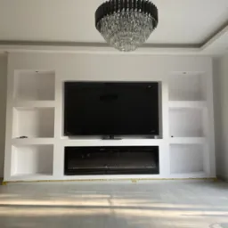 Biała ściana działowa z wnękami, na której zamontowany jest telewizor i elektryczny kominek, pod sufitem wisi kryształowy żyrandol; podłoga zabezpieczona taśmą malarską.