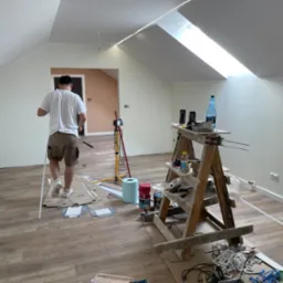 Poddasze w trakcie remontu: świeżo położone panele podłogowe, drabina malarska z narzędziami i farbami, mężczyzna w oddali z listwą przypodłogową, widoczne okno dachowe i oświetlenie LED na suficie.