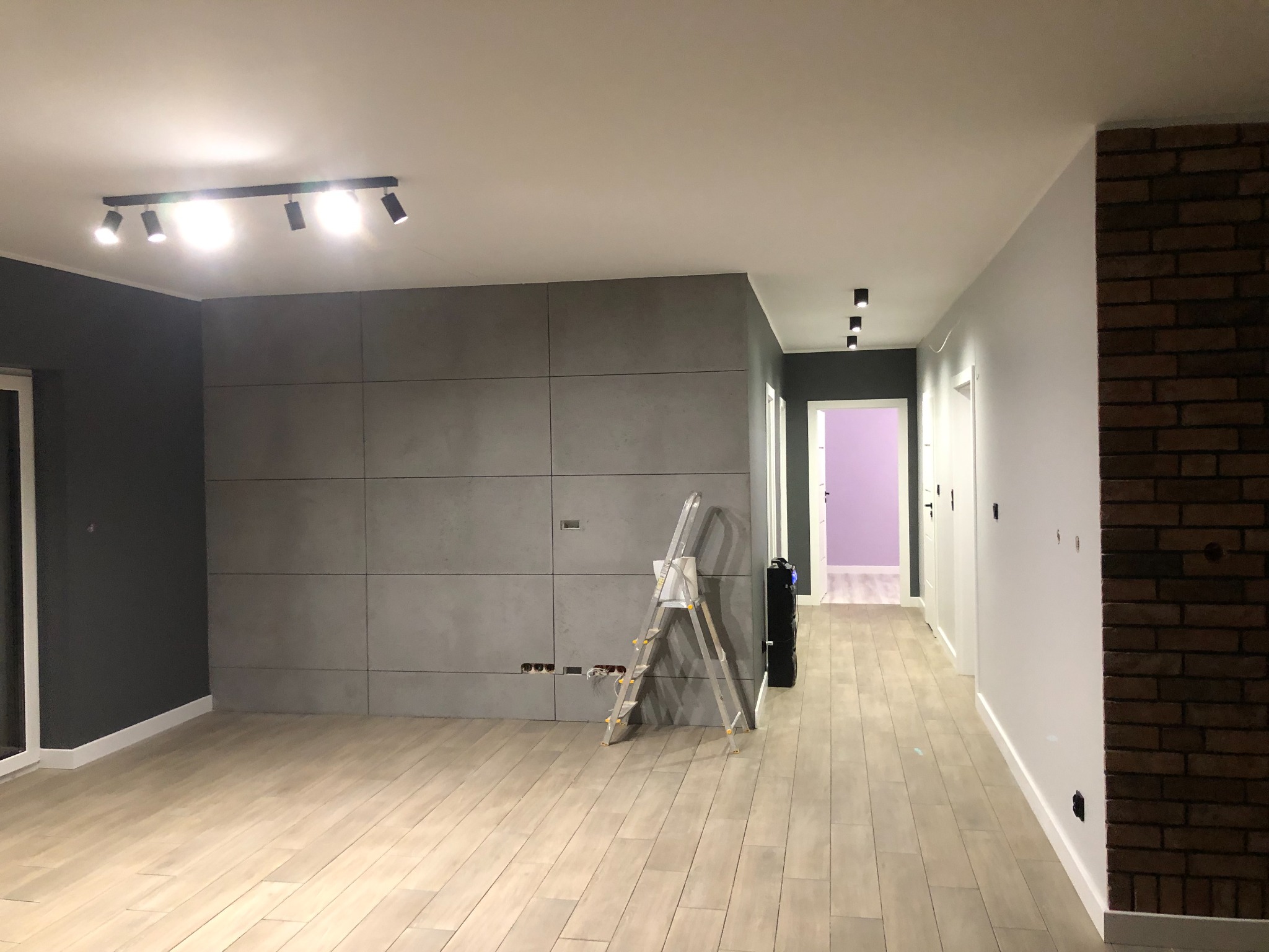 Przestronne wnętrze w trakcie prac wykończeniowych: szare panele ścienne, jasna drewnopodobna podłoga, aluminiowa drabina oparta o ścianę z widocznymi przewodami elektrycznymi, oraz oświetlenie...