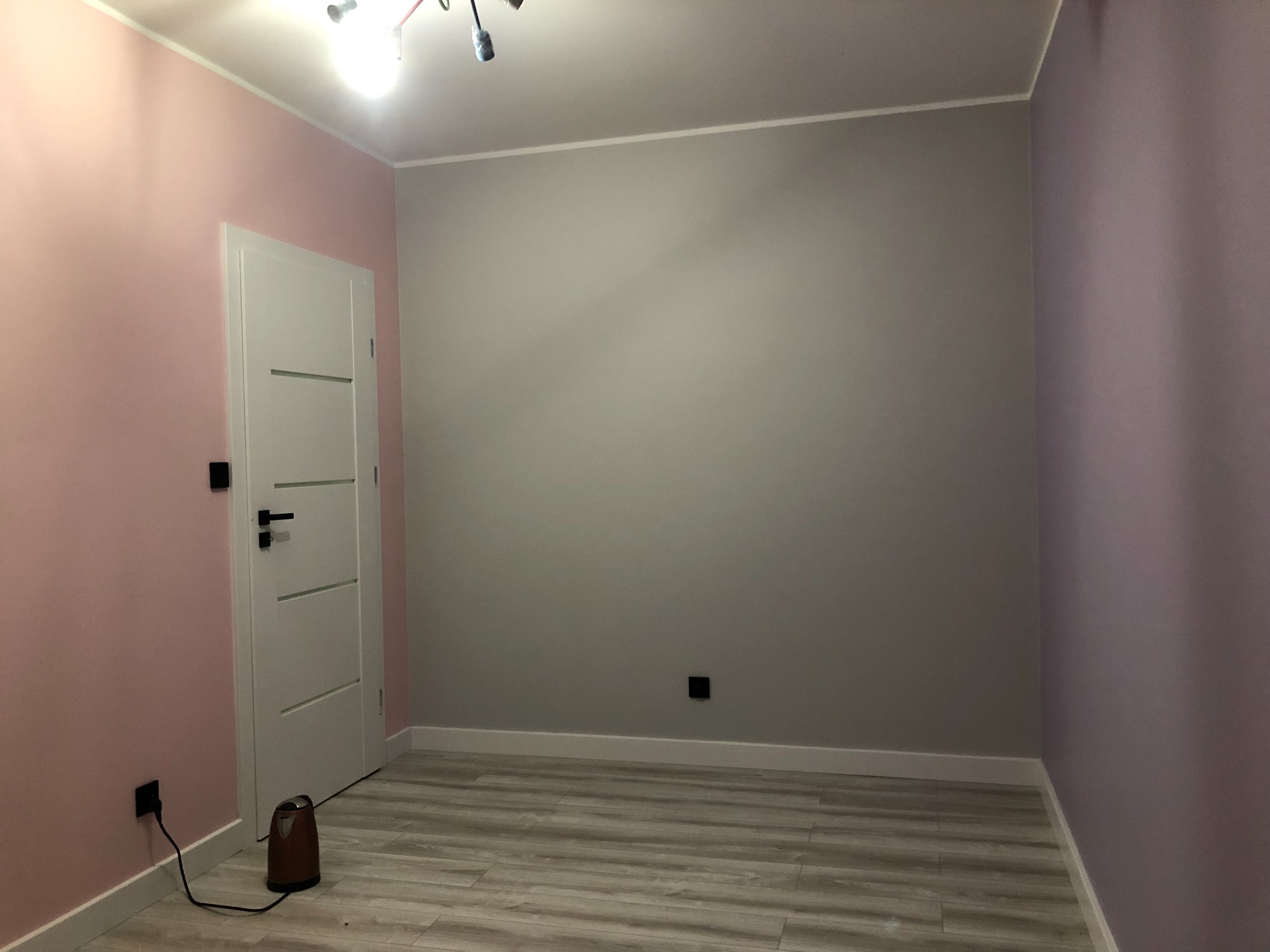 Pomalowane na trzy kolory wnętrze pokoju: ściana różowa przy białych drzwiach z czarną klamką, ściana szara i ściana fioletowa. Na podłodze panele w odcieniach szarości i beżu. Przy ścianie różowej...