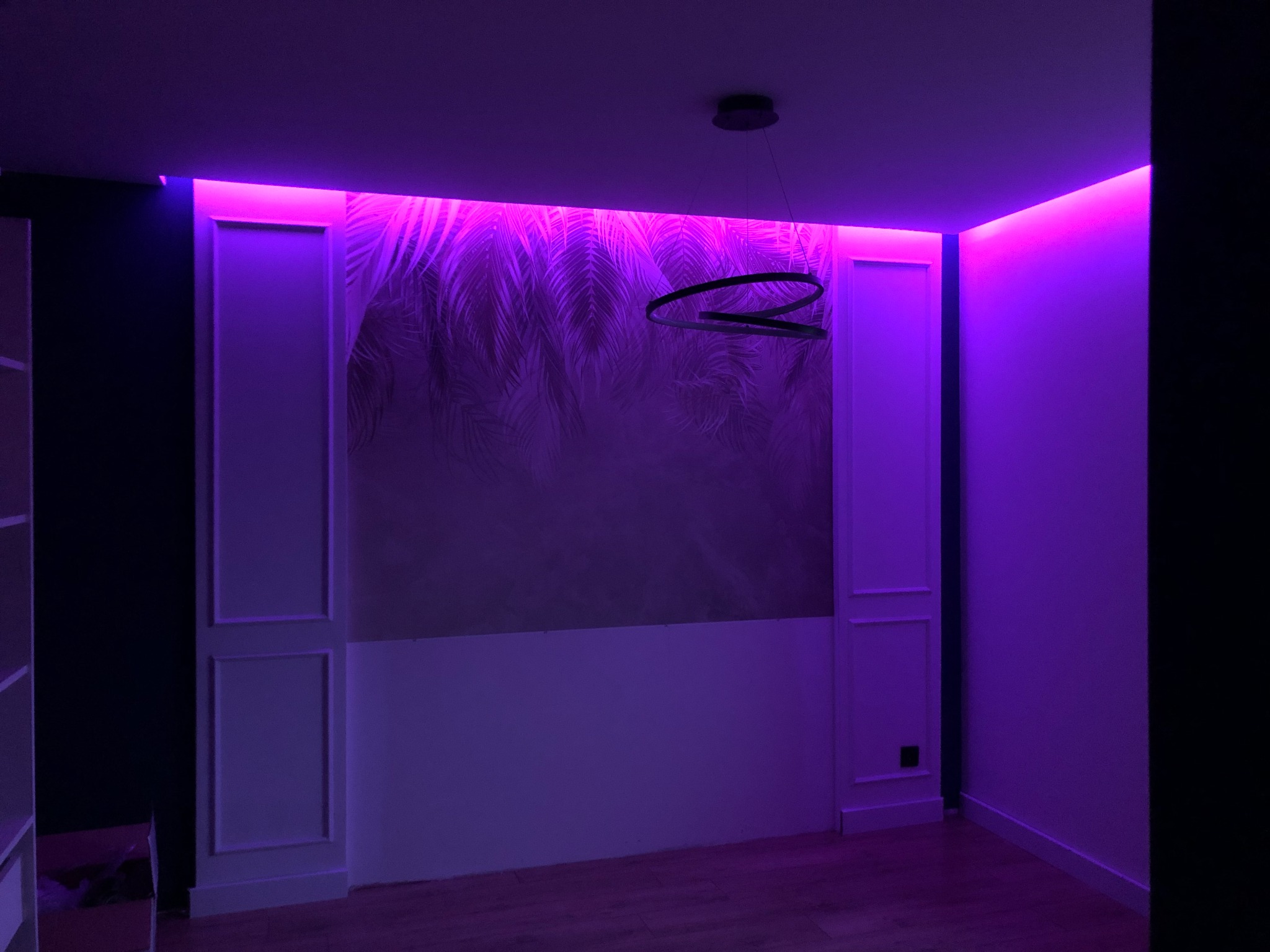 Pokój z dekoracyjnymi panelami ściennymi, tapetą z motywem liści i nowoczesną lampą wiszącą, oświetlony fioletowym światłem LED.