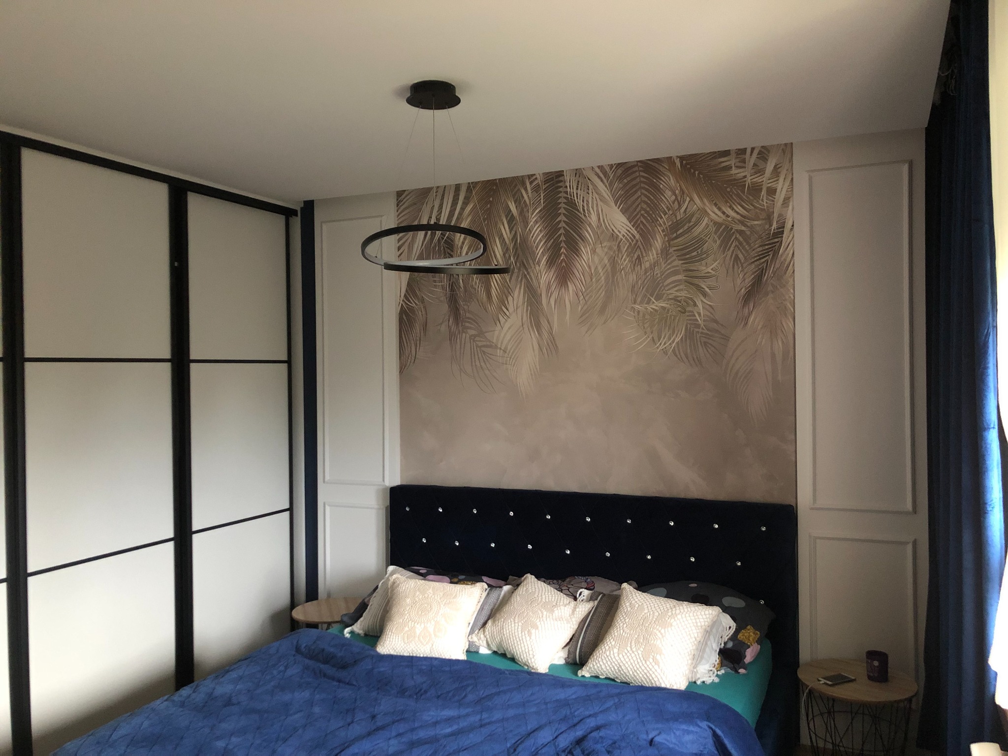 Sypialnia z tapetą w palmy, ciemnoniebieskim łóżkiem z kryształkami, nowoczesną okrągłą lampą i białą szafą przesuwną z czarnymi ramami.