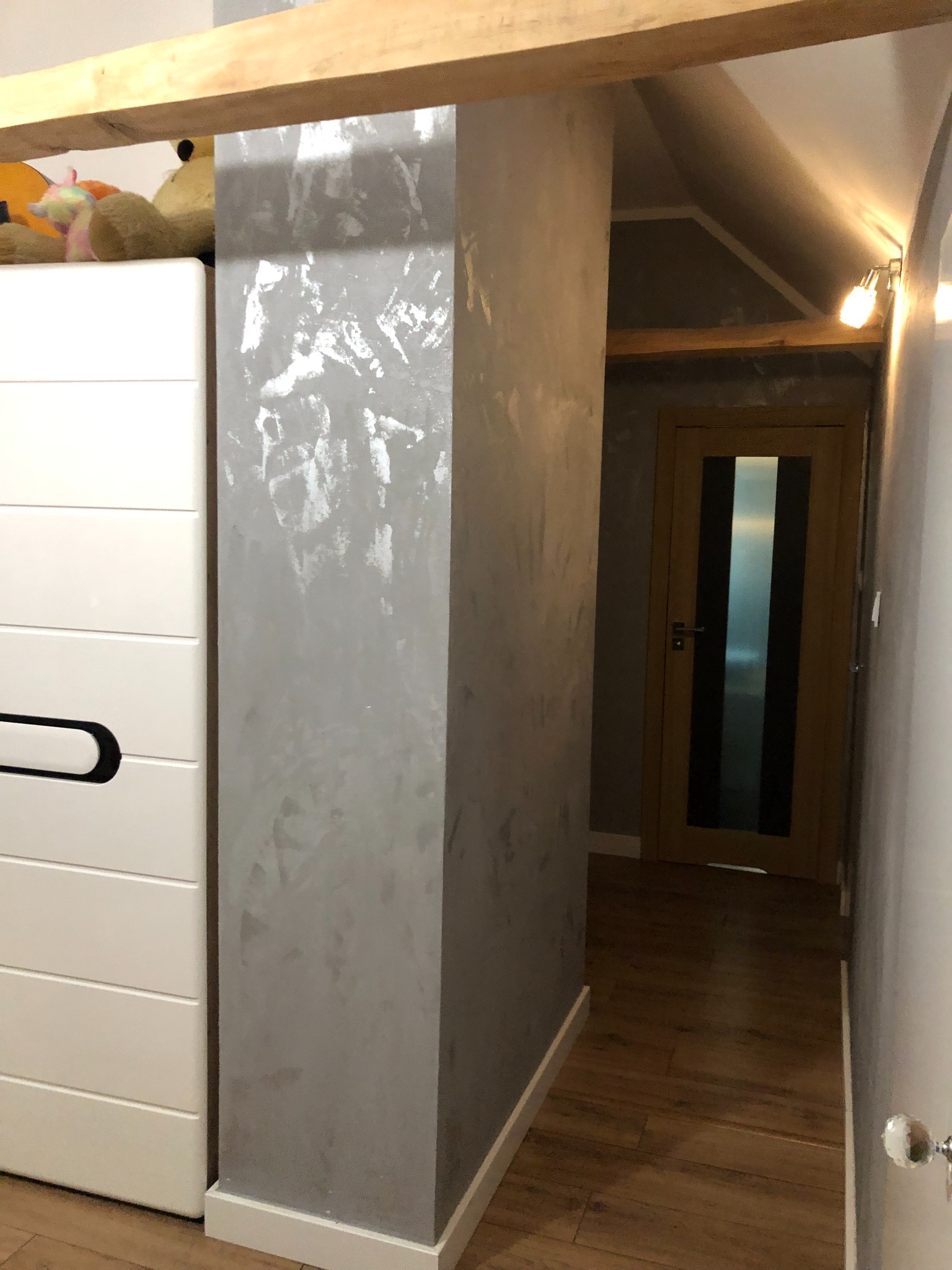 Dekoracyjna kolumna w korytarzu z efektem przetarć, sąsiadująca z białą komodą, drewniana podłoga i drzwi w tle.
