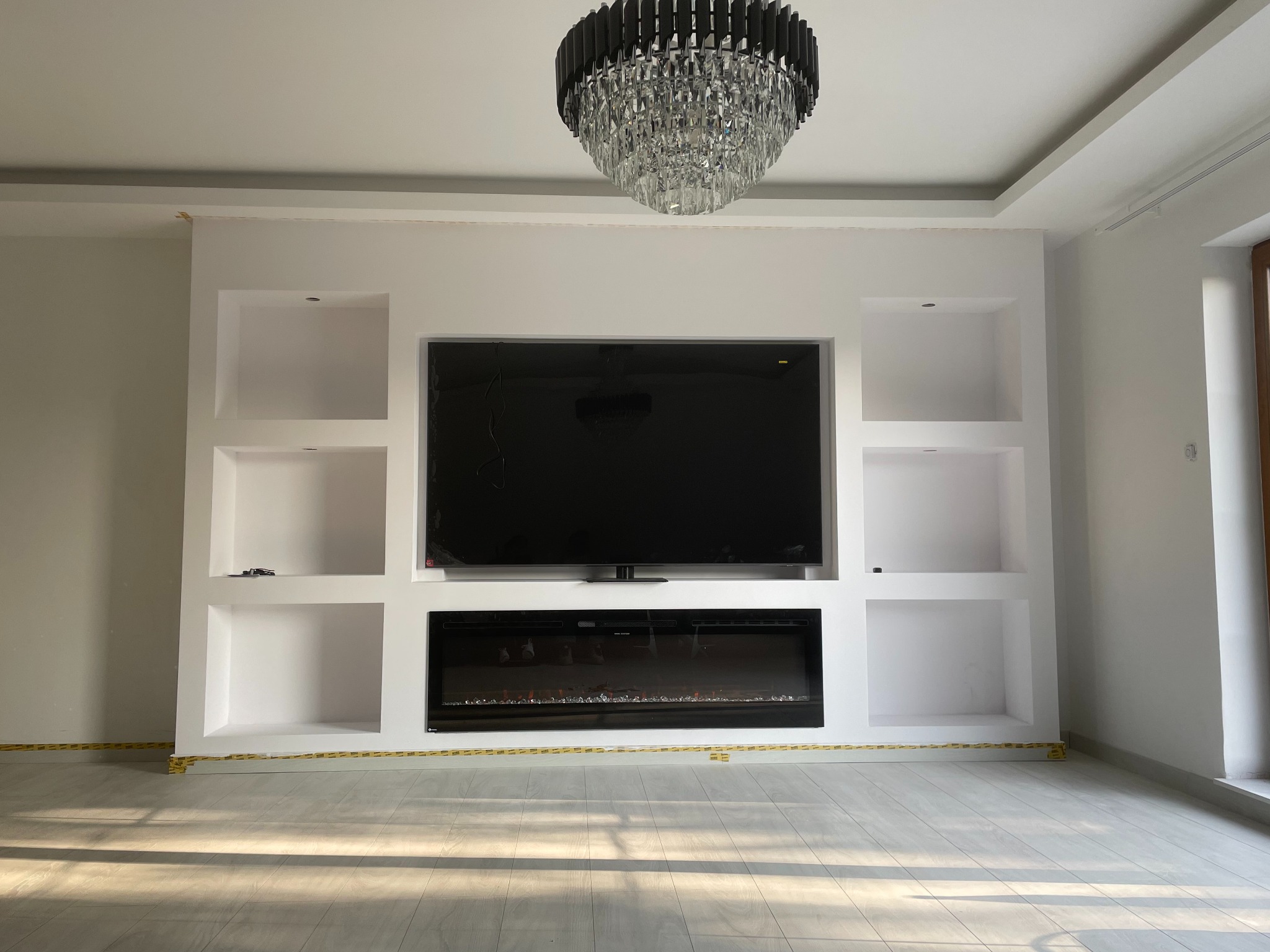 Biała ściana działowa z wnękami, na której zamontowany jest telewizor i elektryczny kominek, pod sufitem wisi kryształowy żyrandol; podłoga zabezpieczona taśmą malarską.