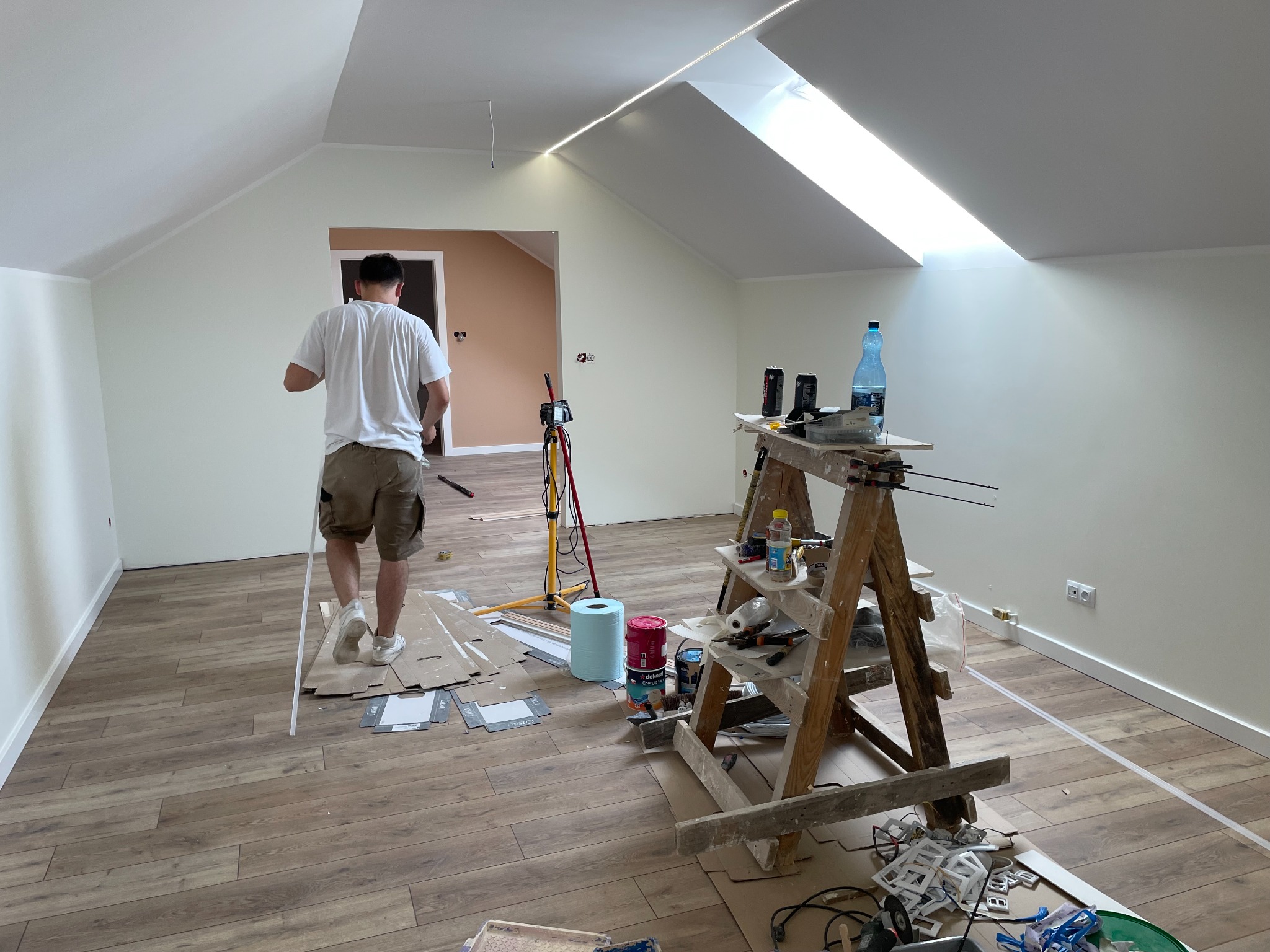 Poddasze w trakcie remontu: świeżo położone panele podłogowe, drabina malarska z narzędziami i farbami, mężczyzna w oddali z listwą przypodłogową, widoczne okno dachowe i oświetlenie LED na suficie.