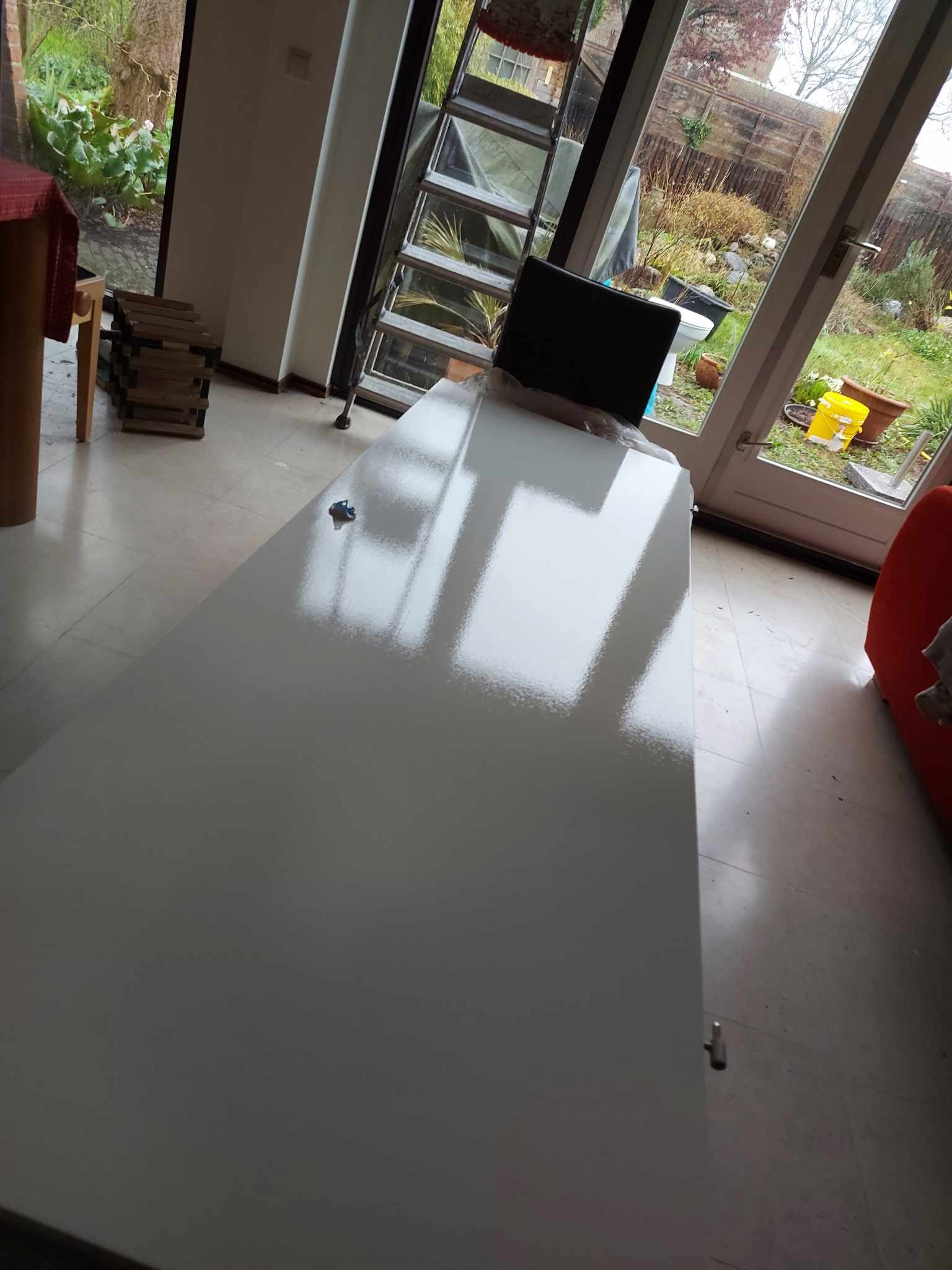 Świeżo pomalowana, biała powierzchnia w pomieszczeniu z widokiem na ogród przez przeszklone drzwi, odbijająca światło z okien.