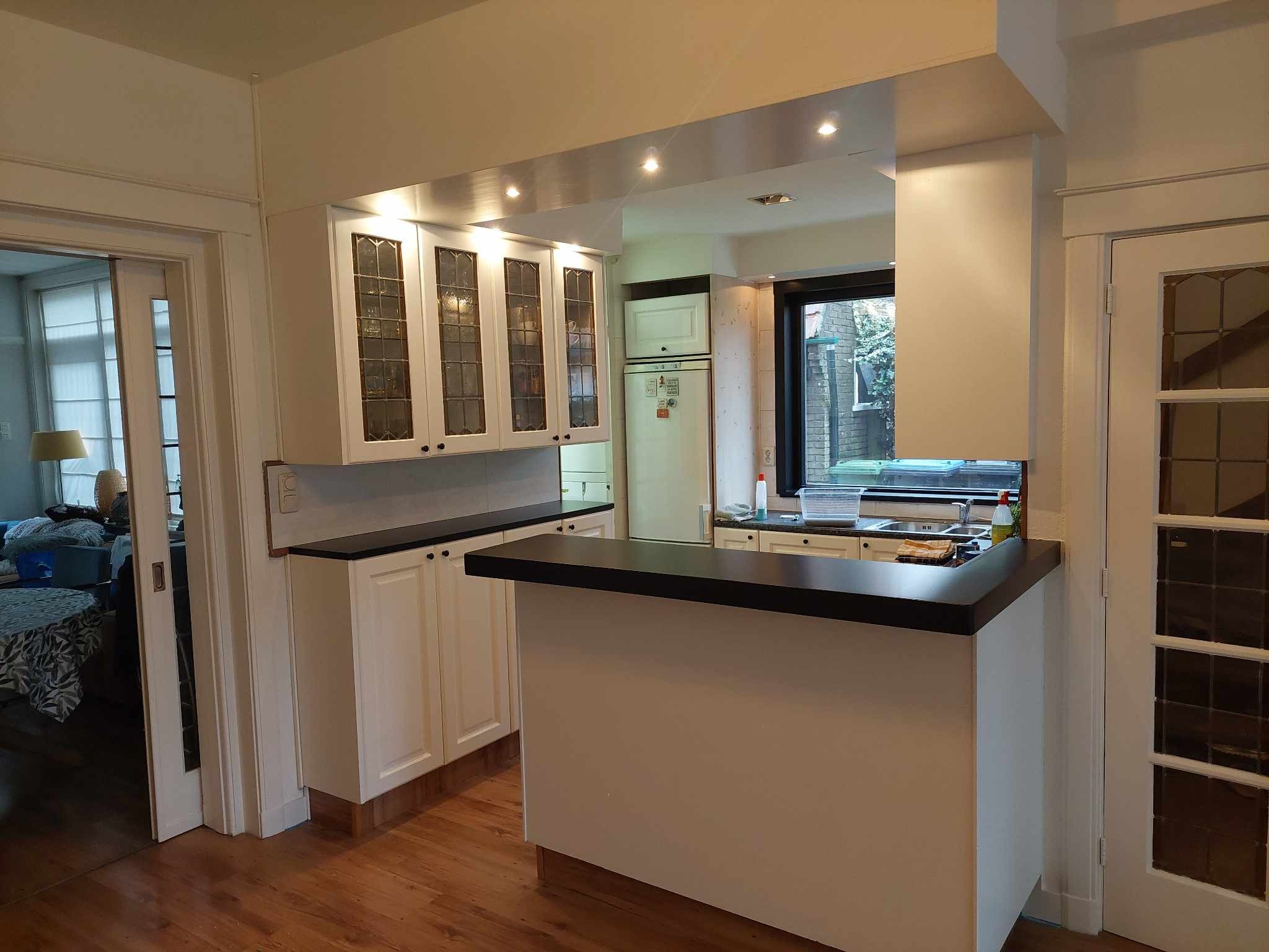 Wykończona kuchnia w stylu retro z białymi szafkami z czarnymi blatami, drewnianą podłogą i oświetleniem punktowym, widok z salonu przez przesuwne drzwi.