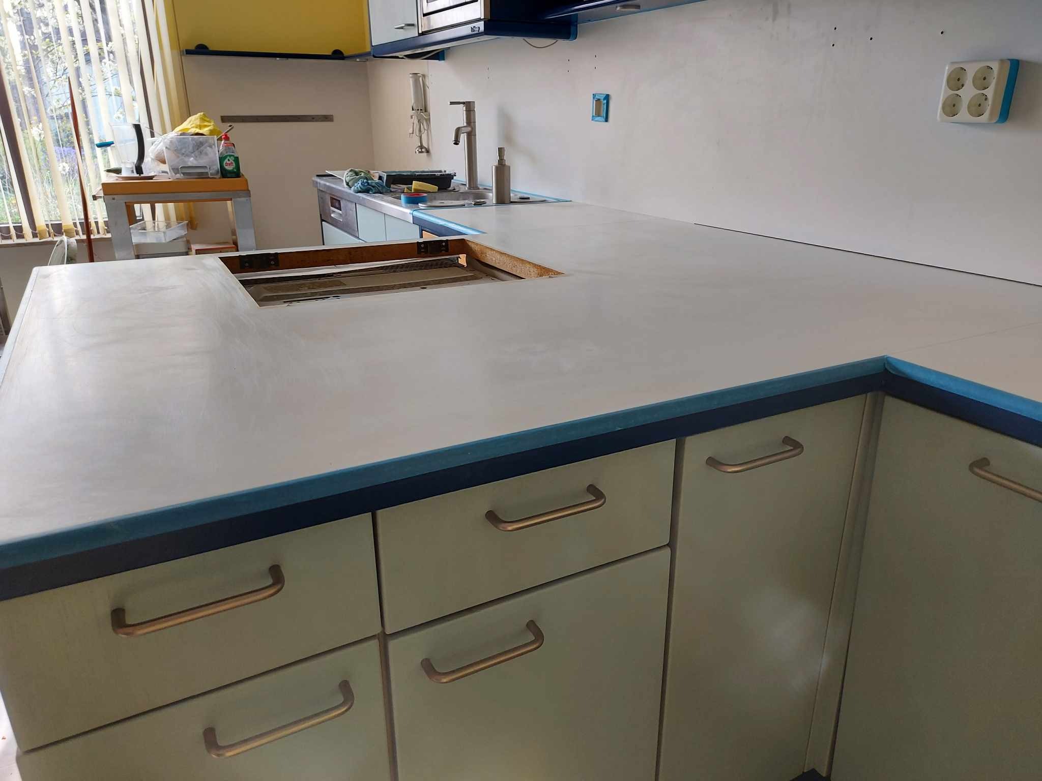 Blat kuchenny z otworem na zlew w trakcie montażu, widoczne szpachlowane ściany i okno z żaluzjami w tle.