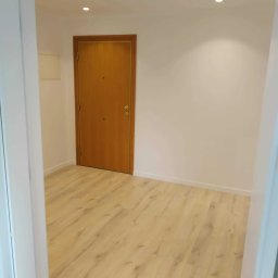 Lux-mig - Wnętrze pustego, odświeżonego pokoju z panelami podłogowymi w odcieniach beżu i brązu, widok na drewniane drzwi wejściowe i białe ściany oświetlone punktowymi lampami sufitowymi.