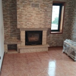 Lux-mig - Wykończony kominek z obudową z płytek imitujących kamień, z boczną półką, w pokoju z terakotową podłogą i oknem w czarnej ramie, widok na fragment wnętrza po remoncie.