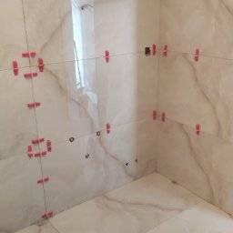 Lux-mig - Ściany i podłoga łazienki w trakcie układania płytek imitujących marmur, z widocznymi czerwonymi klipsami poziomującymi i sznurkami, otwory na instalacje widoczne na ścianie.