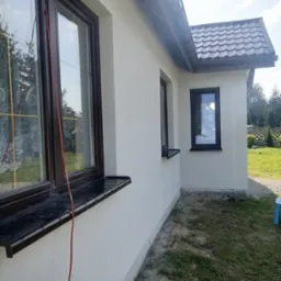 Świeżo pomalowana, jasna elewacja domu jednorodzinnego z brązowymi oknami i ciemną dach&oacute;wką, widok z boku, na trawniku leży czerwony kabel.