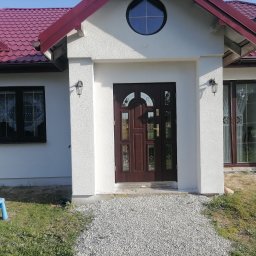 Lux-mig - Biały dom z bordowym dachem i brązowymi drzwiami wejściowymi, okrągłe okno nad wejściem, ścieżka z białego żwiru prowadząca do drzwi.